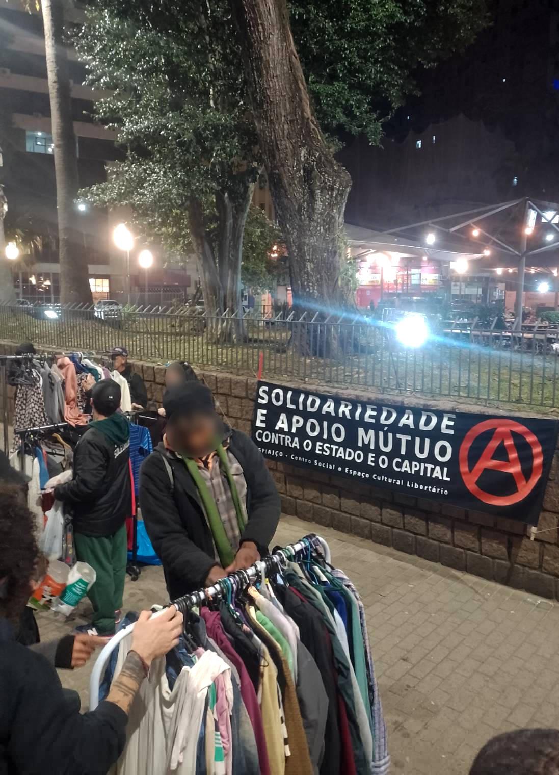 Foto. Em primeiro plano pessoas olhando roupas penduradas em três araras. Ao fundo  uma faixa de tecido preto pendurada em um muro de pedra. Na faixa está escrito em letras garrafais brancas: "Solidariedade e apoio mútuo contra o Estado e o Capital." Abaixo em letras menores, está escrito: "Espaço - Centro Social e Espaço Cultural Libertário". E ao lado um grande A vermelho dentro de um círculo.